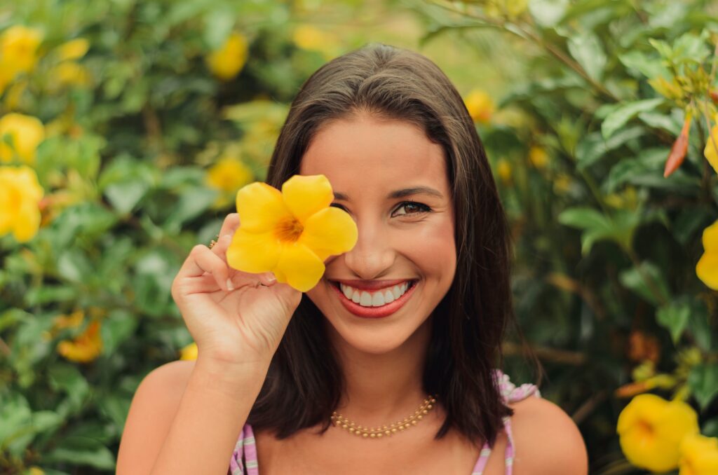 Woman smiling at the camera and holding a yellow flower over her eye